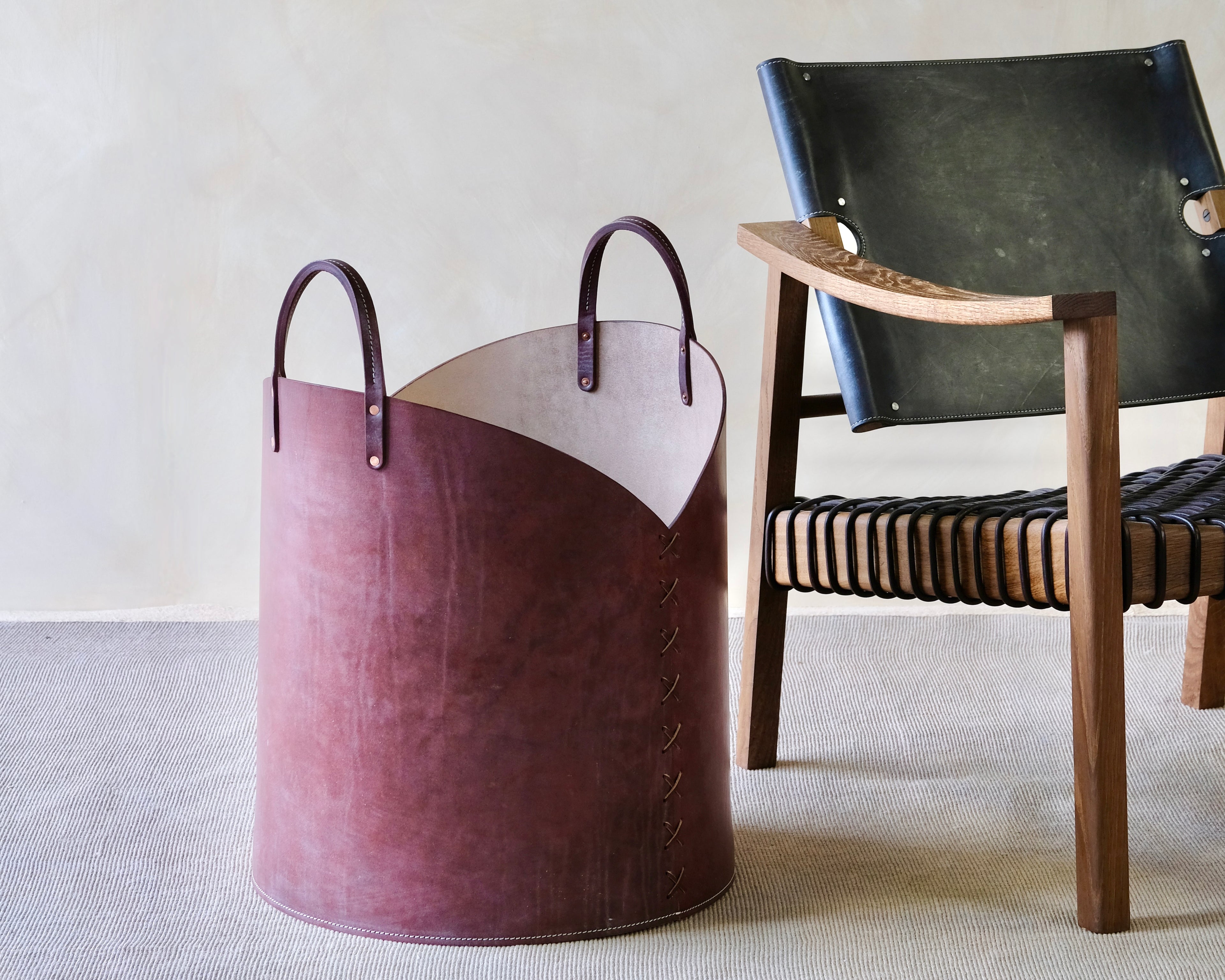 Burgundy leather basket with handles next to a black leather chair on a light gray floor.