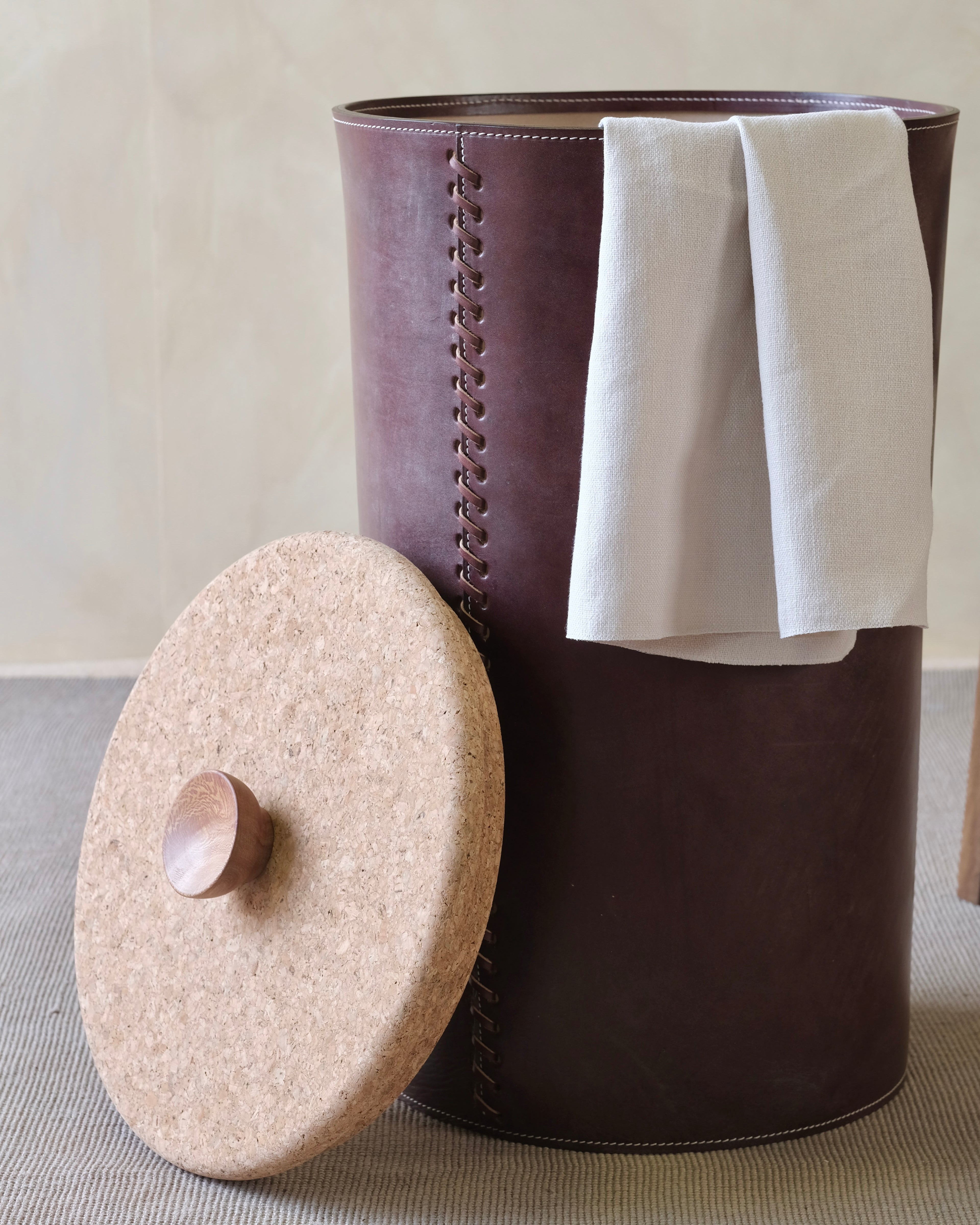 Burgundy brown leather laundry basket with a white towel and beige lid on a neutral background