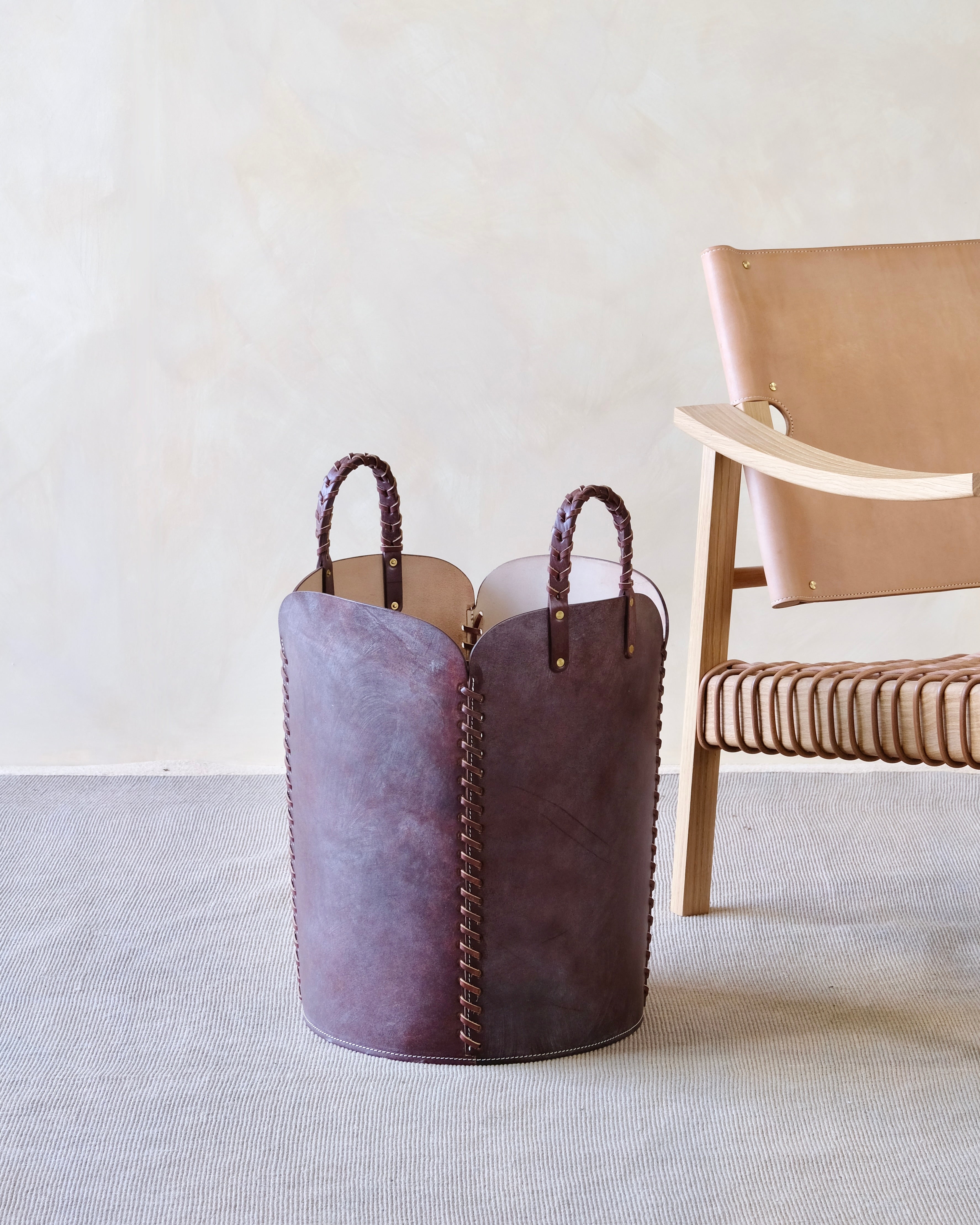 Burgundy leather basket with handles on a light gray floor with a beige chair in the background.
