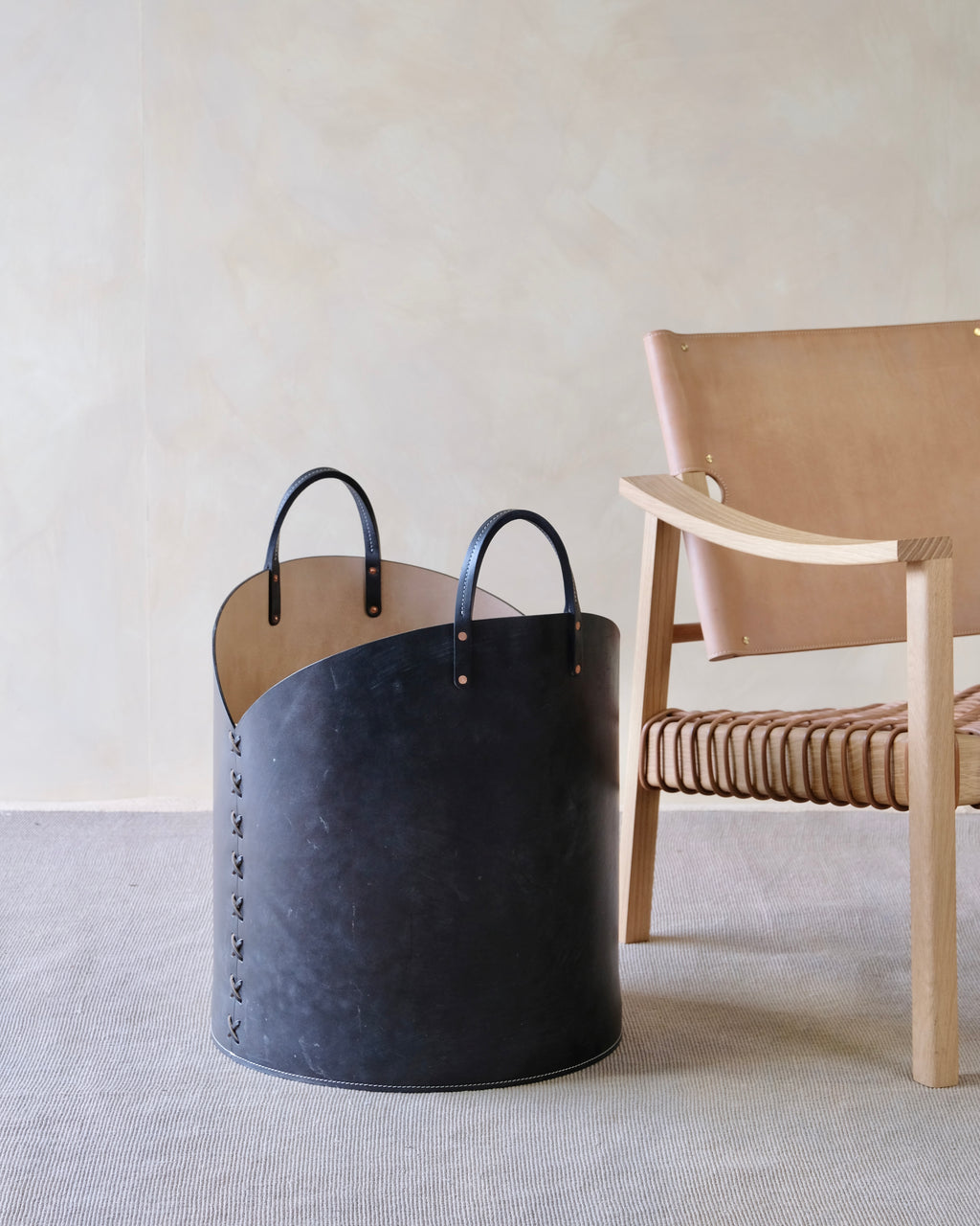 Black leather basket with handles on a light gray surface next to a wooden chair.