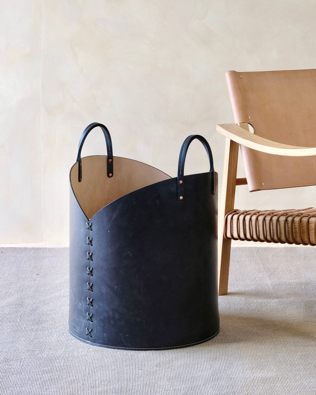 Black leather basket with handles on a light gray surface with a beige wall and wooden chair in the background.