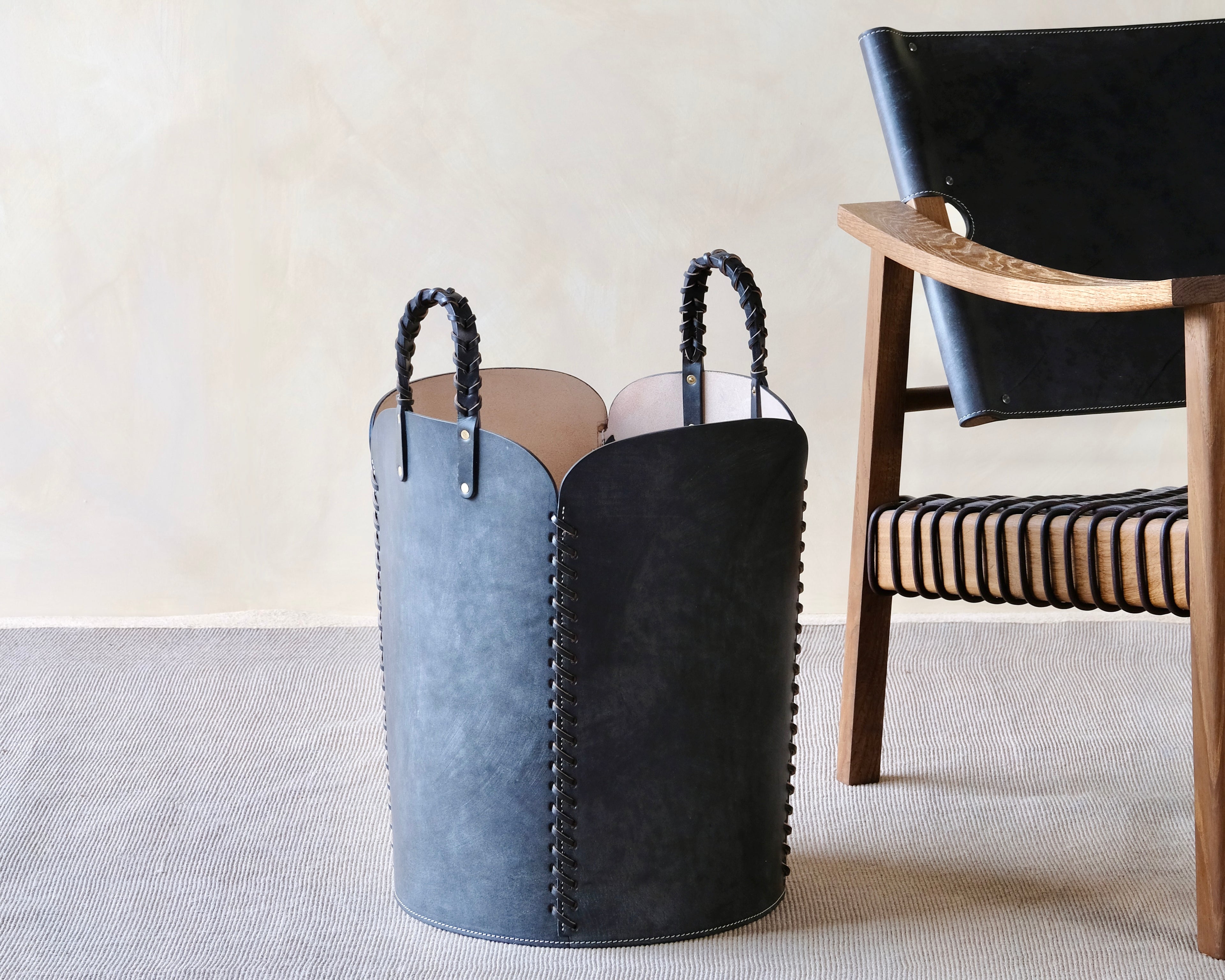Black leather basket with handles on a light gray floor next to a wooden chair.