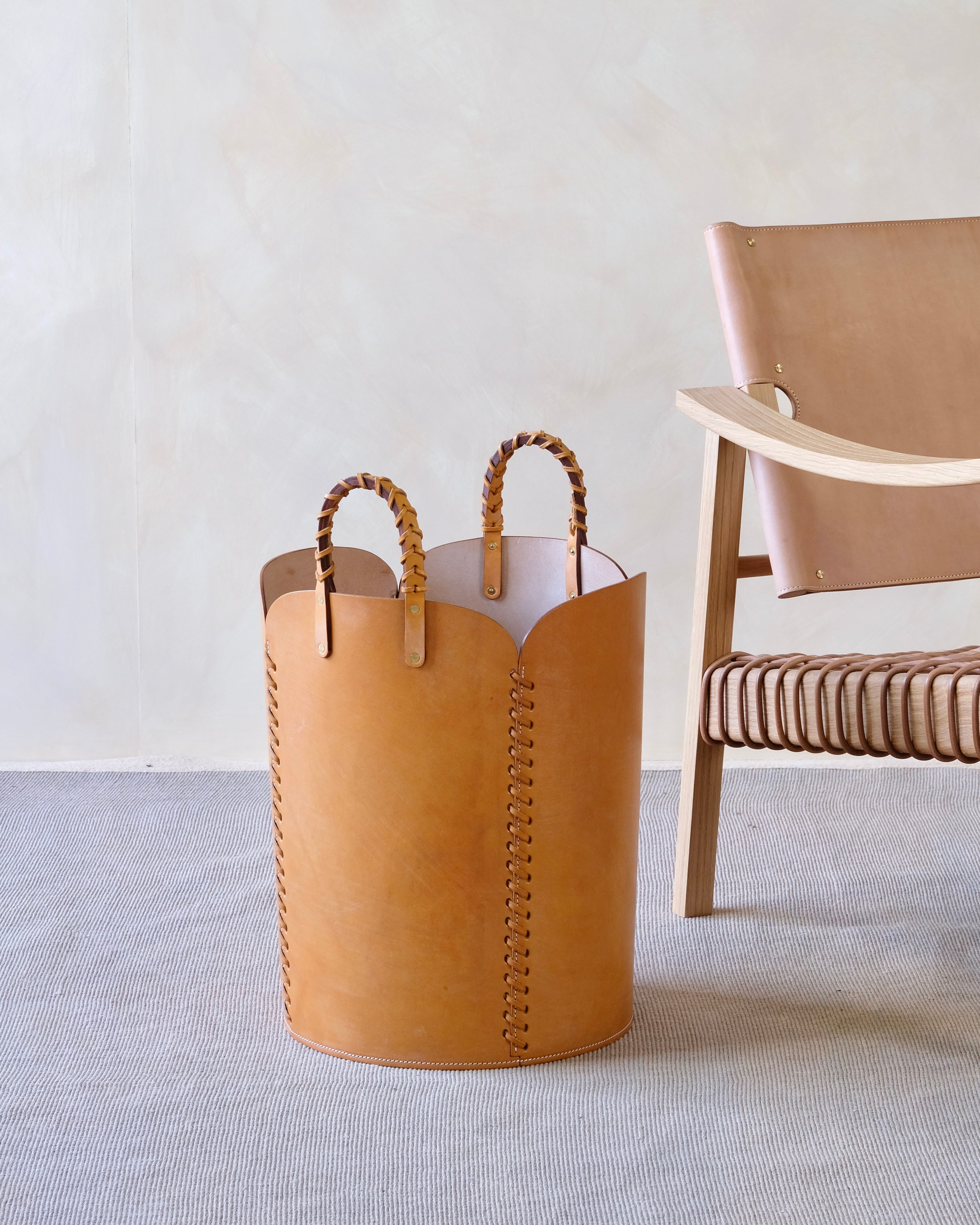 Tan leather basket with woven handles on a light gray floor next to a wooden chair.
