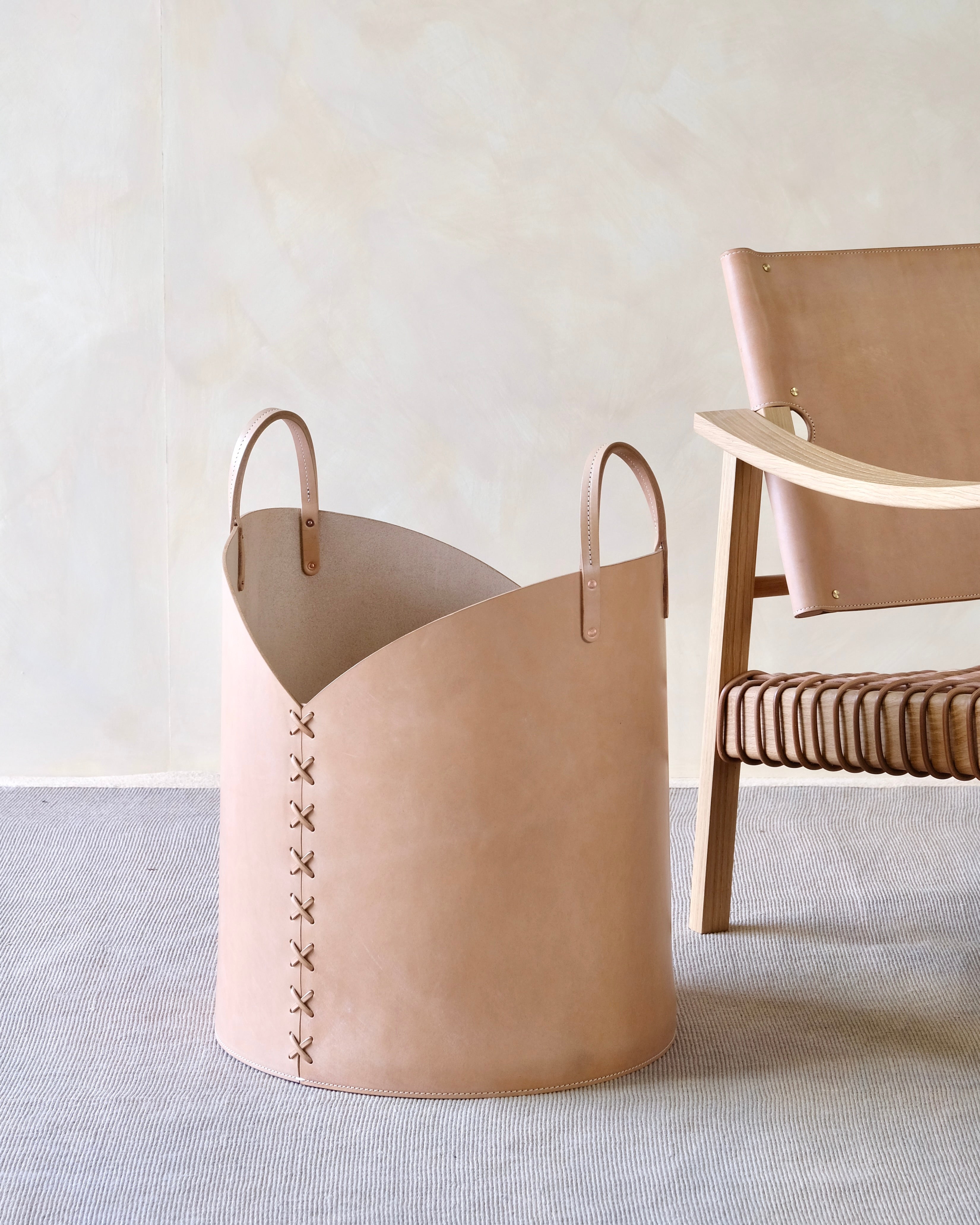 Beige leather basket with decorative stitching on a light gray surface with a wooden chair in the background.