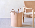 Beige leather basket with handles next to a wooden chair on a light gray surface.