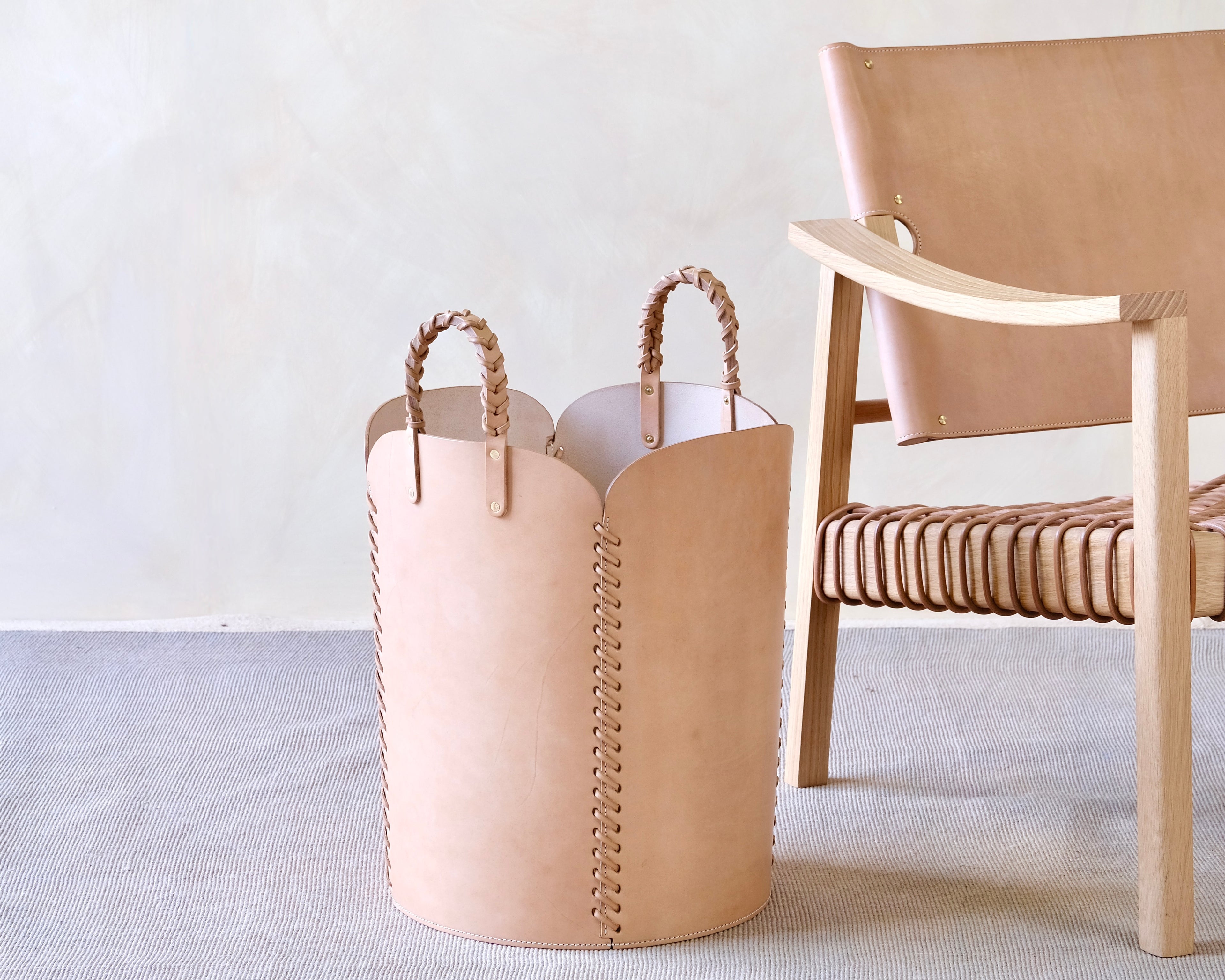 Beige leather basket with handles next to a wooden chair on a light gray surface.