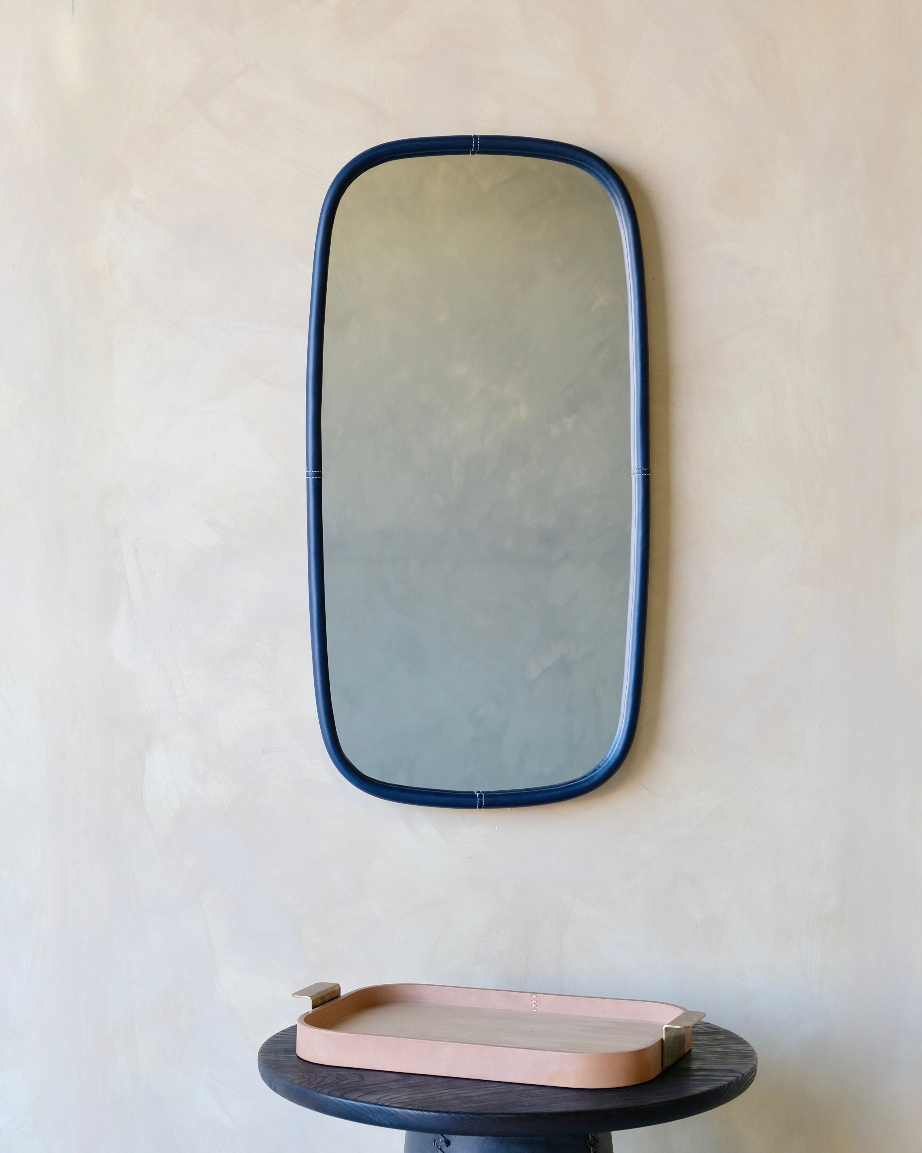 Rectangular mirror with blue frame on a beige wall above a round dark surface with a pink tray.